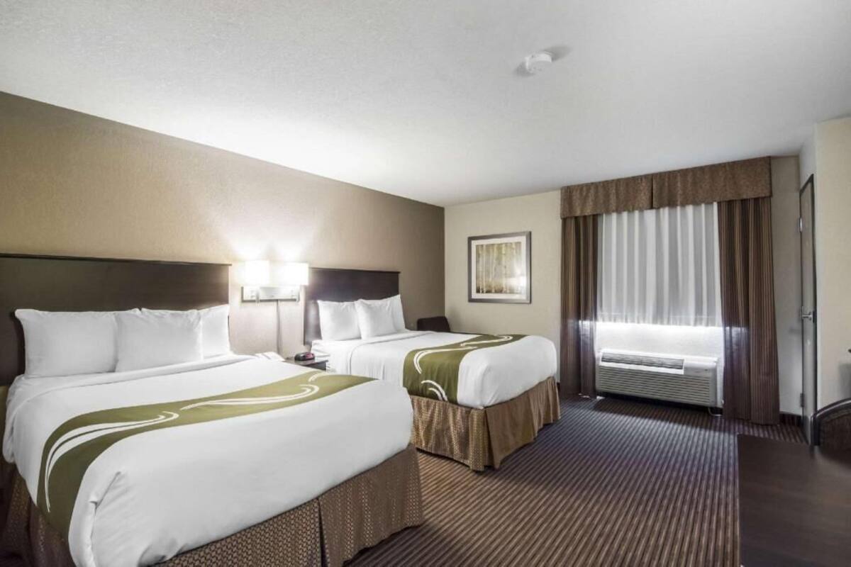 A hotel room with two beds, a large window with curtains, a wall-mounted air conditioner, and a framed picture on the wall.
