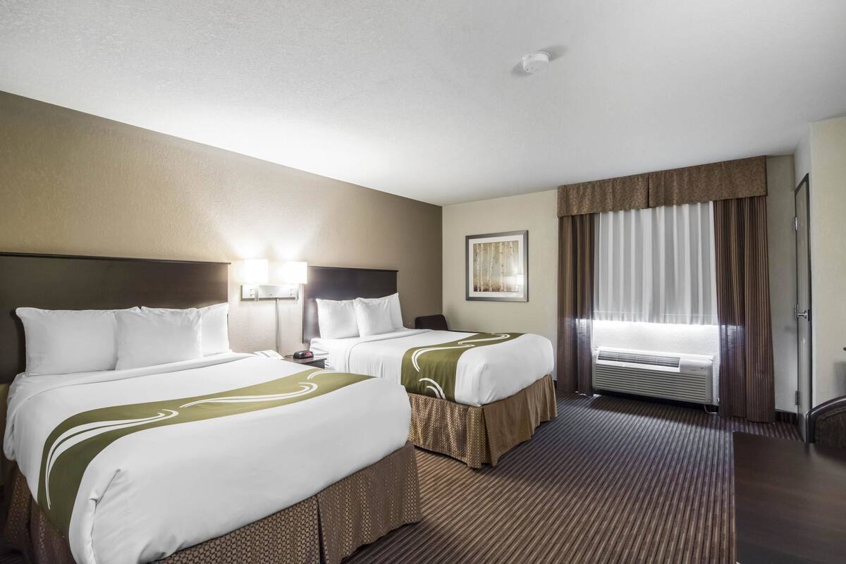 A hotel room with two beds, a large window with curtains, a wall-mounted air conditioner, and a framed picture on the wall.