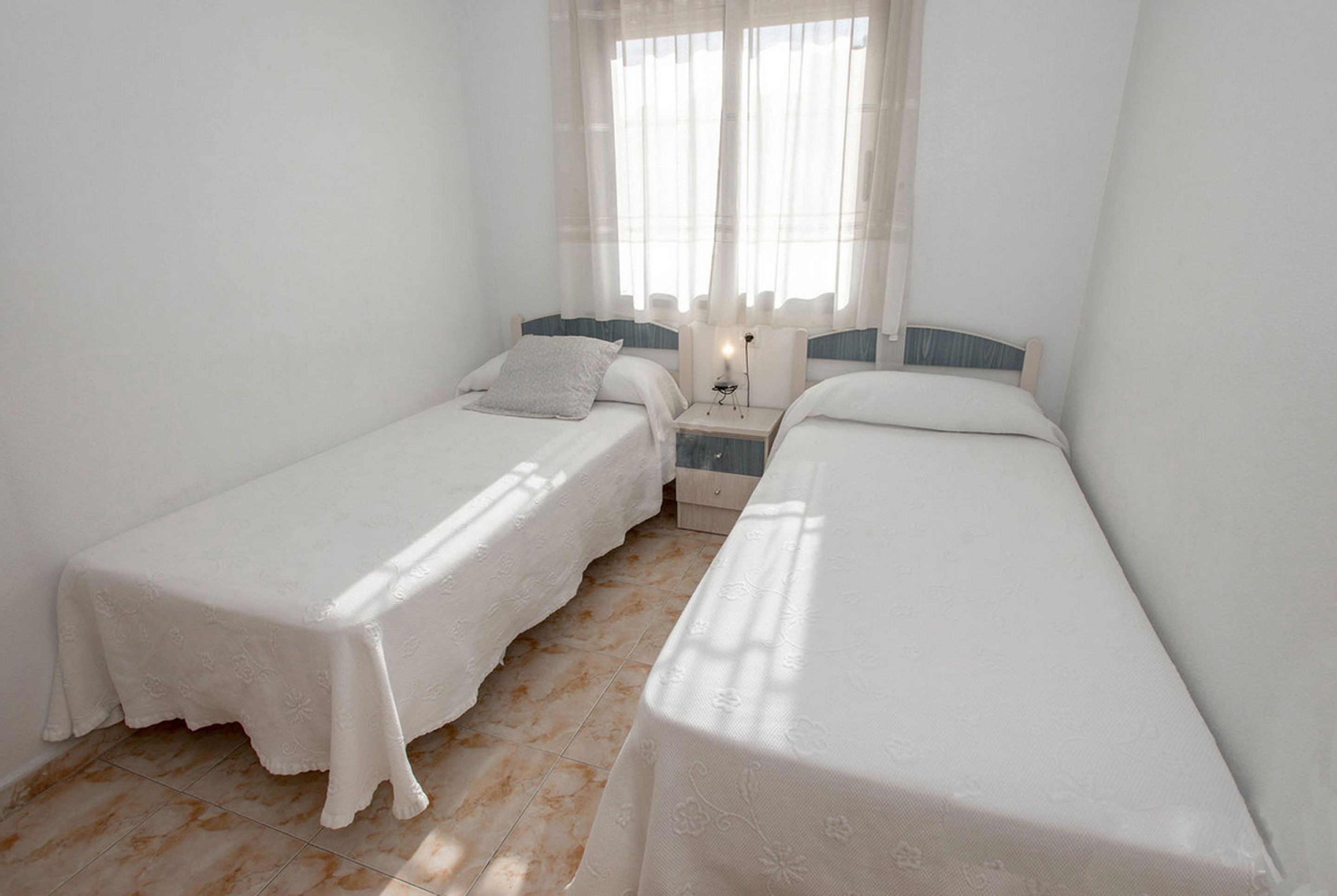 Una habitación con dos camas individuales, una mesita de noche pequeña con lámpara y cortinas traslúcidas.
