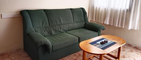 A green sofa, a wooden coffee table with a placemat and a plate of fruit, and a window with white curtains.