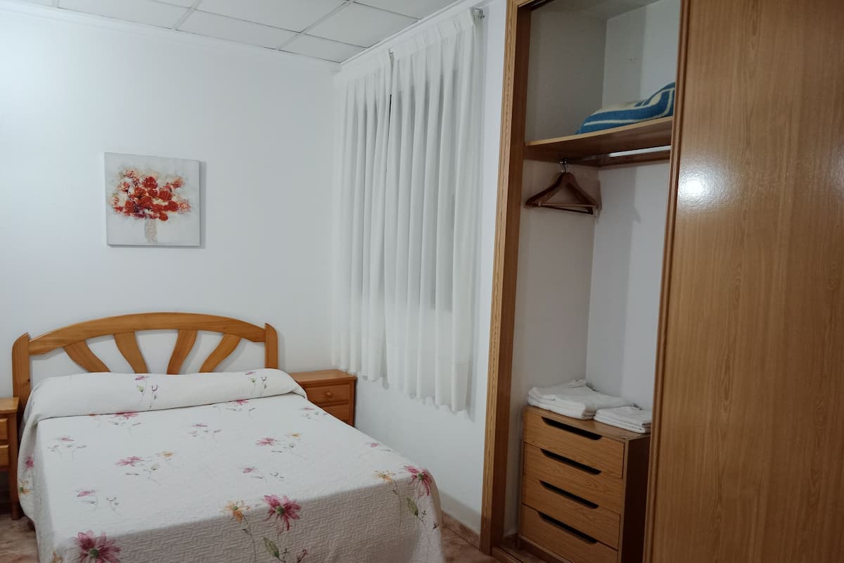 A bedroom with a wooden bed, a nightstand, a wardrobe, and a window with white curtains.