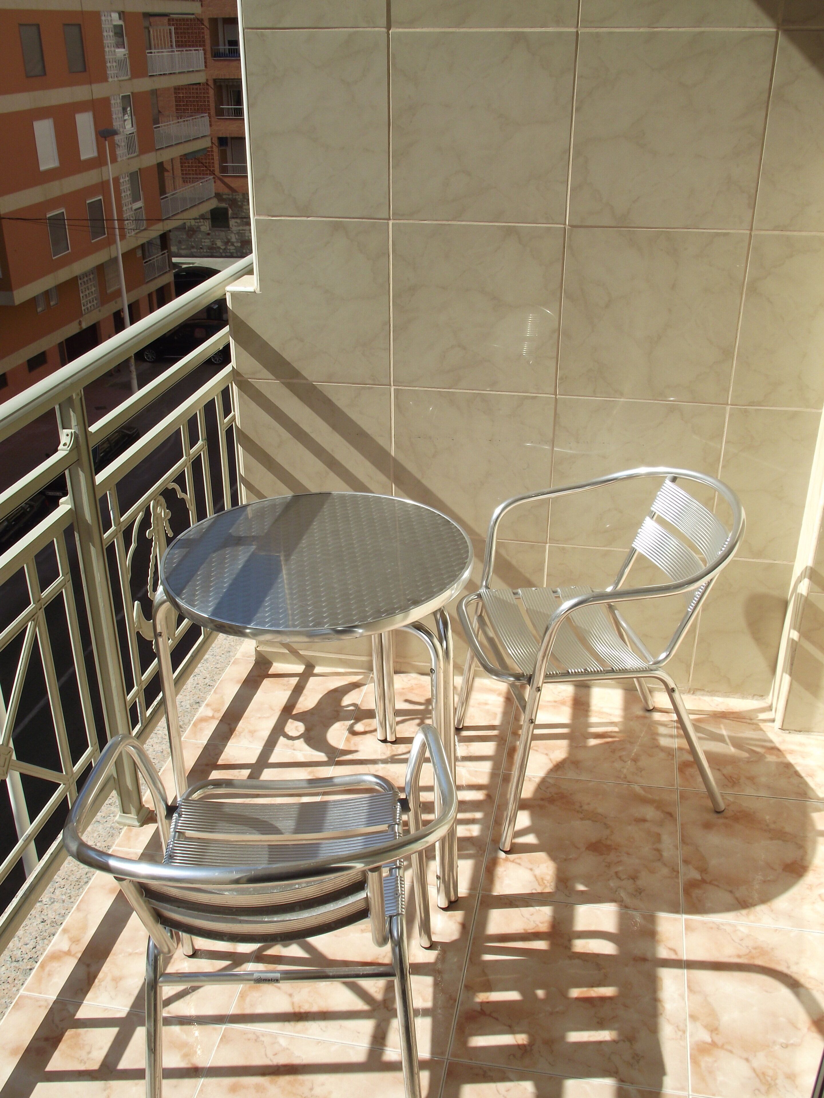 A balcony with a round table and two metal chairs, a tiled floor, and a view of a building in the background.