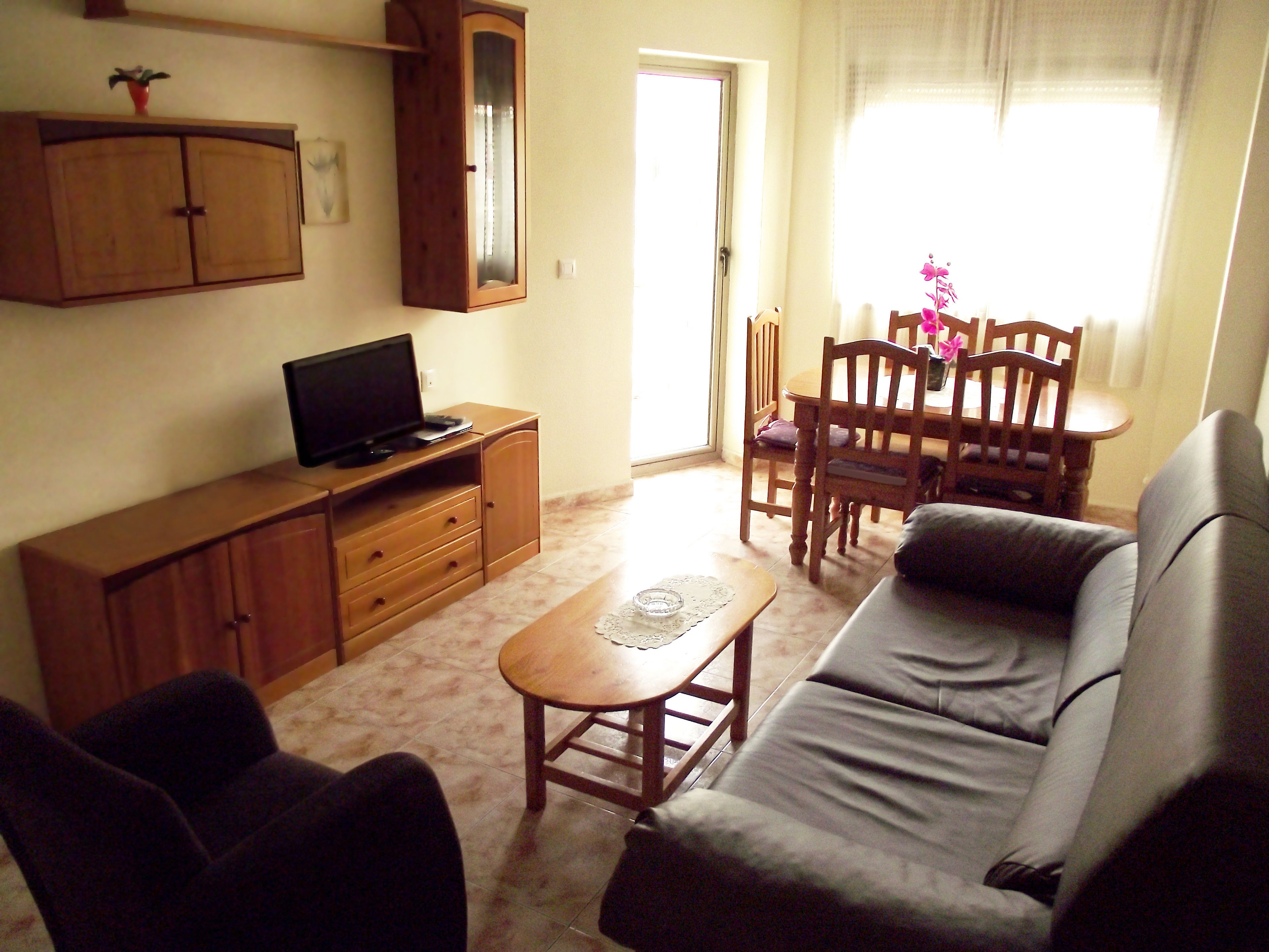 Un salon comprenant un canapé, un téléviseur posé sur un meuble en bois, une table à manger avec des chaises et une fenêtre avec des rideaux.