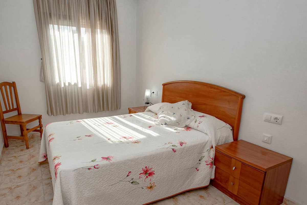 A bedroom with a bed, a wooden nightstand, a chair, and a window with curtains.