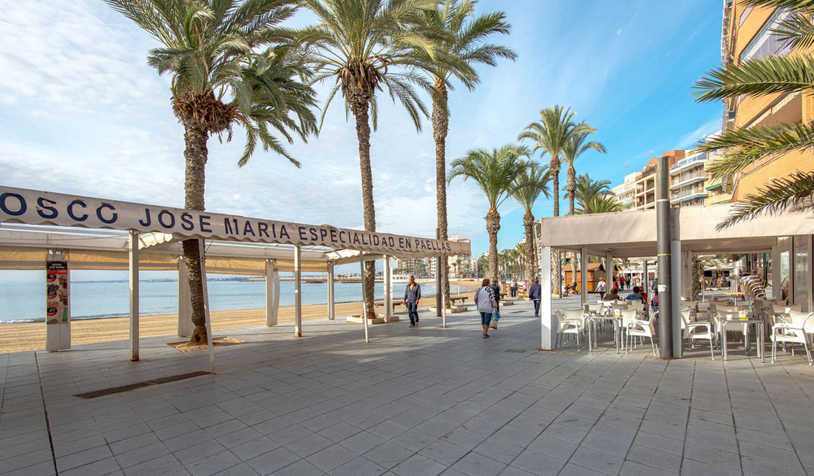 Une promenade en bord de mer bordée de palmiers, une enseigne indiquant « OSCC JOSE MARIA, spécialité : paellas », et des espaces de restauration en plein air.