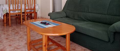 A living room with a green sofa, wooden chairs, a small wooden table with a placemat, and a wall-mounted picture of potted plants.