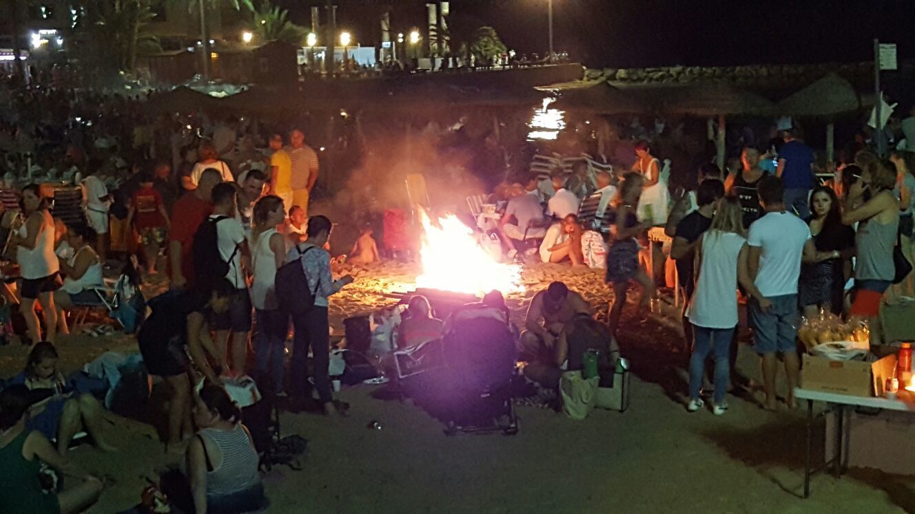 Reunión al aire libre concurrida alrededor de una fogata central, con gente sentada y de pie en grupos.