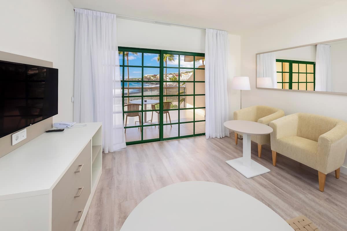 Una habitación de hotel moderna con una televisión de pantalla plana, un sofá, una pequeña mesa redonda y un balcón con vistas al mar.