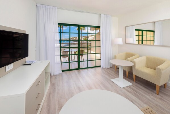 Una habitación de hotel moderna con una televisión de pantalla plana, un sofá, una pequeña mesa redonda y un balcón con vistas al mar.