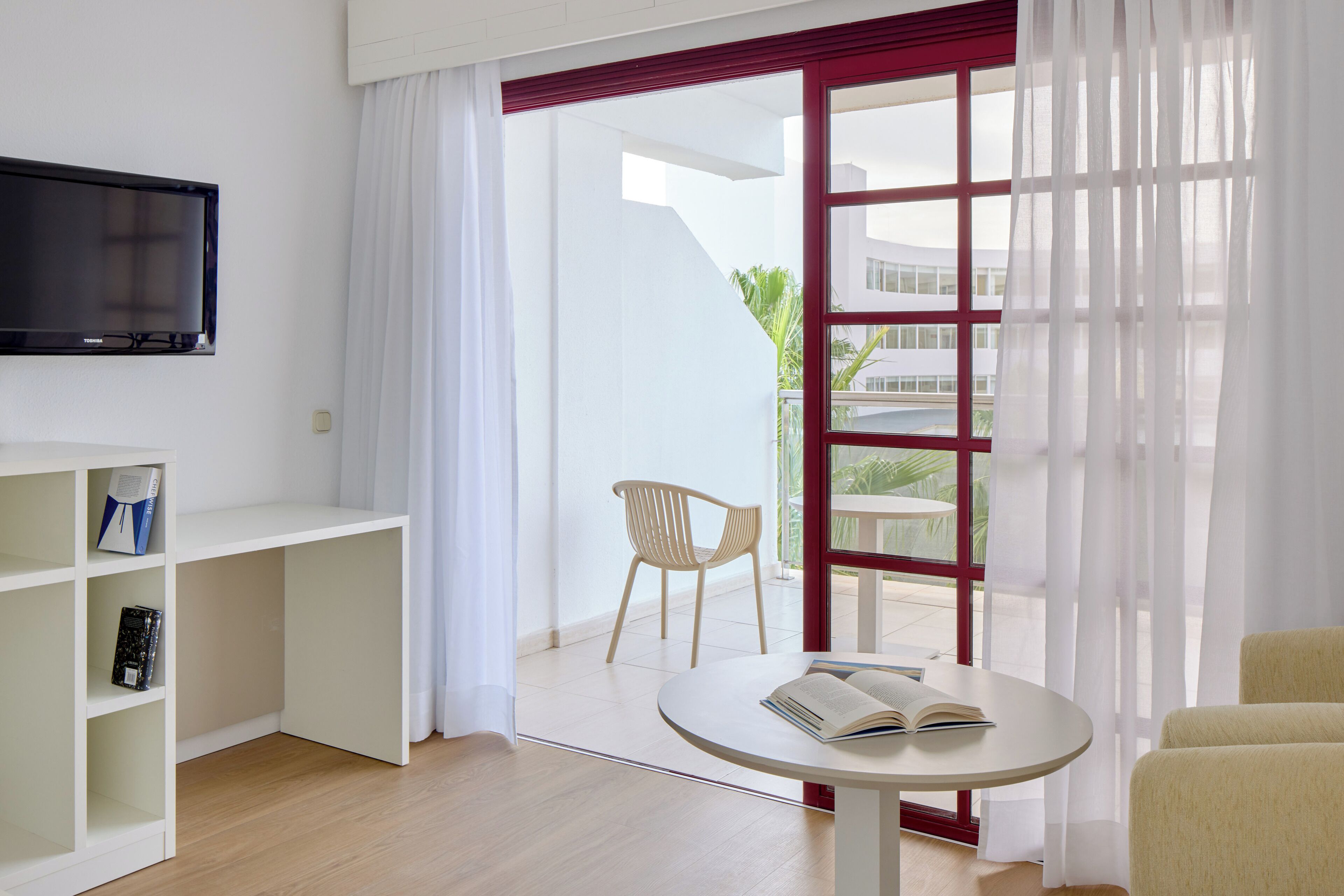 A modern room with a flat-screen TV, a white shelving unit, a round table with a book, a chair, and a view of a building through a glass door.