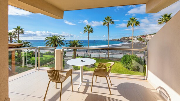 A balcony with a table and chairs, overlooking a beach with palm trees and a clear sky.