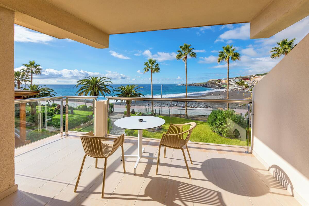 Un balcón con mesa y sillas, con vista a una playa con palmeras y cielo despejado.