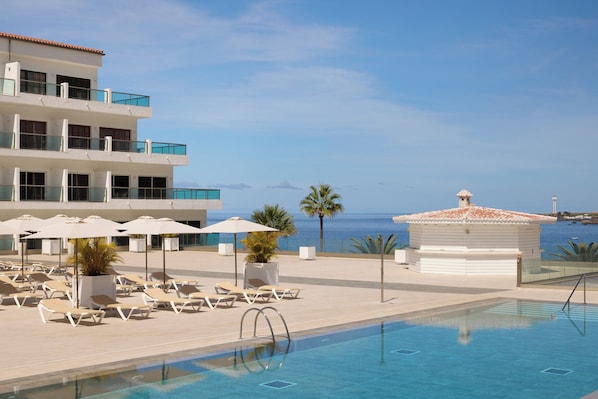 A pool area with lounge chairs and umbrellas, a building with balconies, and a view of the sea.