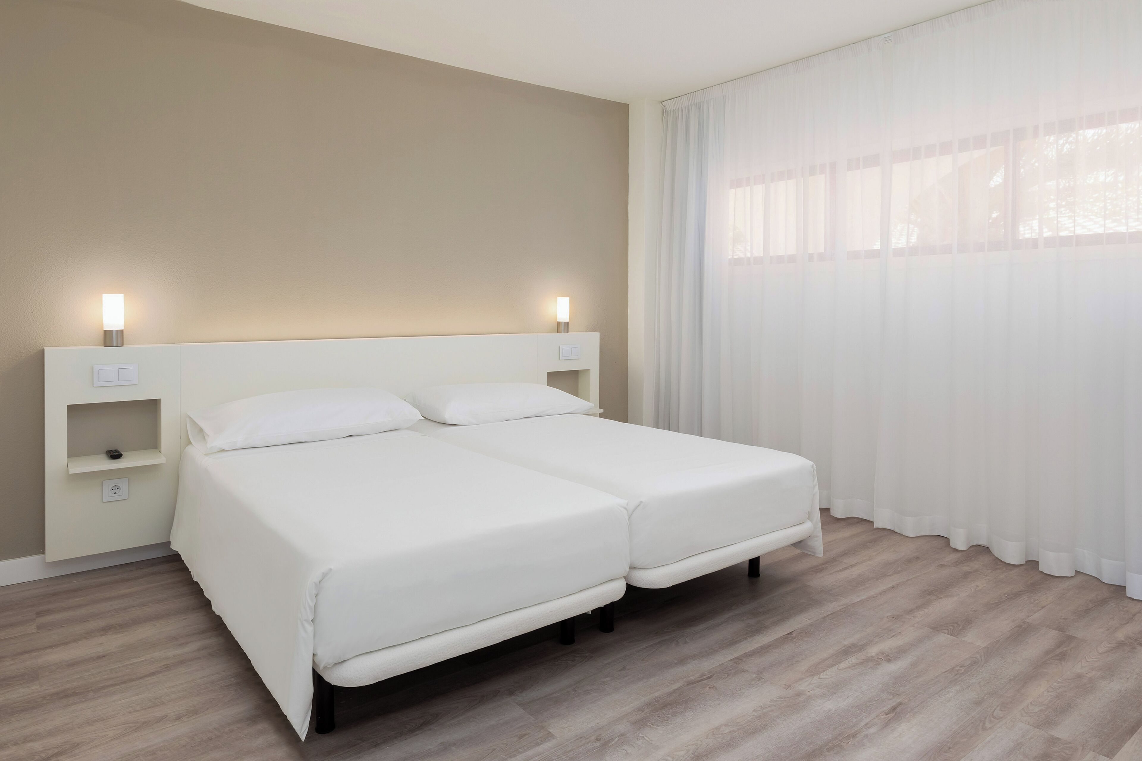 Uma cama de casal com cabeceira branca, uma mesinha de cabeceira com abajur e uma janela com cortinas finas.