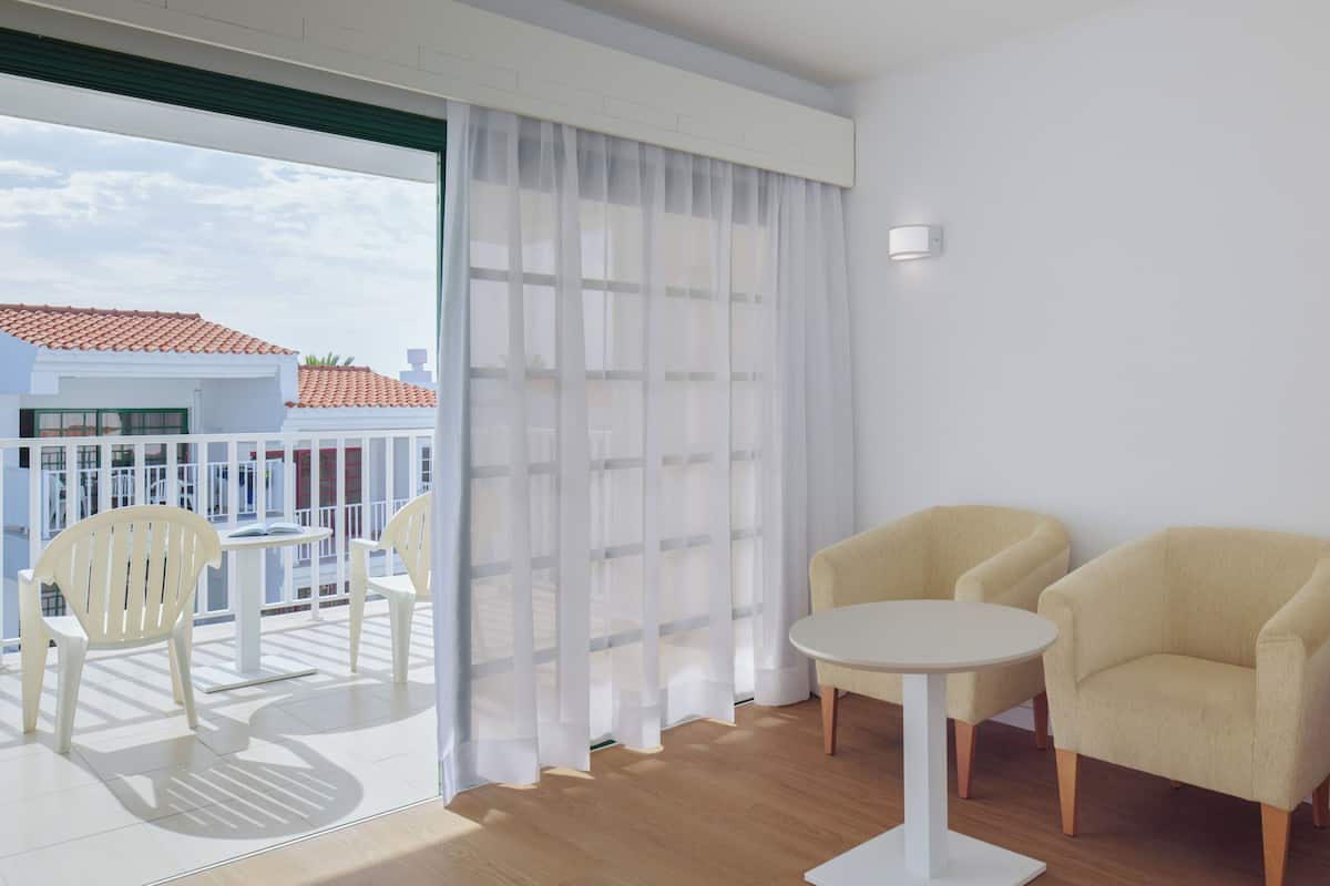 Un balcón con sillas y una mesa blancos, una pared blanca con un interruptor de luz, y vistas de edificios con tejados rojos.
