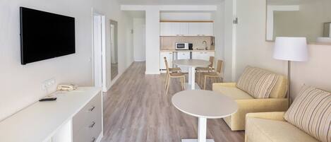 Ein modernes Hotelzimmer mit Flachbildfernseher, Schreibtisch mit Schubladen, rundem Tisch, Sofa, Küchenzeile mit Schränken und Mikrowelle sowie einem großen Spiegel.
