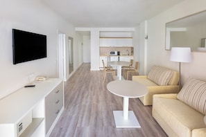A modern hotel room with a flat-screen TV, a desk with drawers, a round table, a sofa, a kitchen area with cabinets and a microwave, and a large mirror.