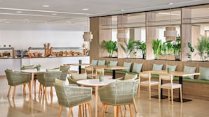 A modern dining area with wicker chairs and tables, a kitchen in the background, and a large window with plants.
