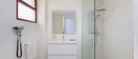 Un baño moderno con ducha envuelta en vidrio, tocador blanco con espejo y secador de pelo en la pared.