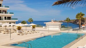 A pool area with lounge chairs and umbrellas, a small building, and a view of the sea.