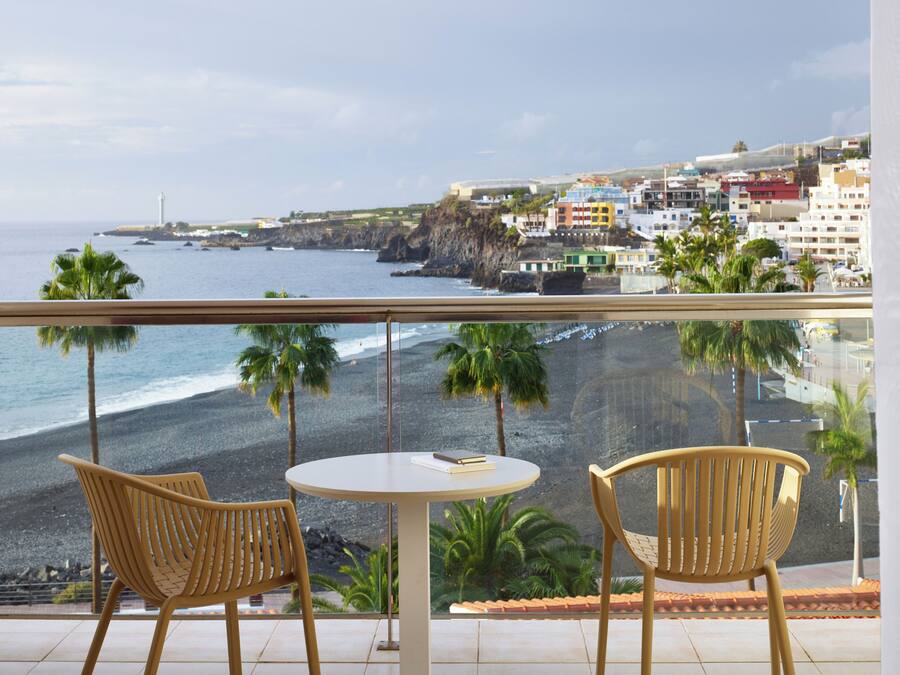 Un balcón con dos sillas de mimbre y una pequeña mesa redonda, con vista a una playa de costa rocosa y un pueblo al fondo.