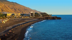 A coastal hotel with multiple floors, a beach in the foreground, and a rocky cliff in the background.