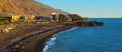 Un hotel frente al mar con varios pisos, una playa en primer plano y un acantilado rocoso al fondo.
