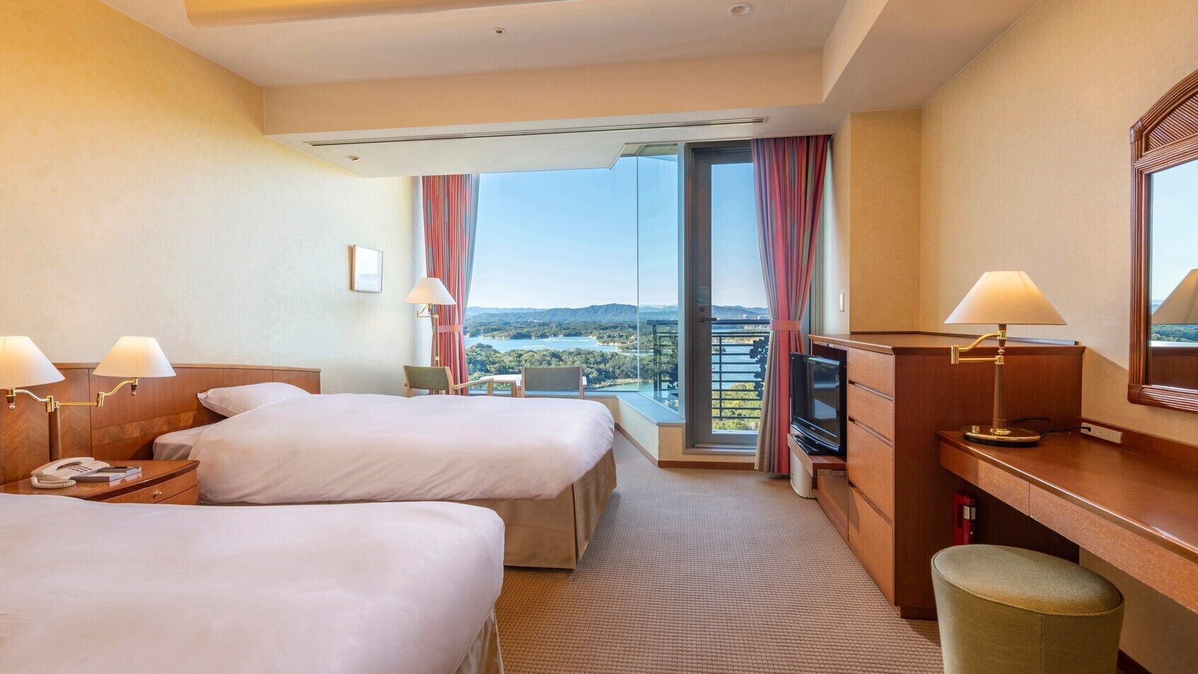 Ein Hotelzimmer mit zwei Betten, einem Fernseher, einem Schreibtisch mit Lampe und Blick auf eine Landschaft.