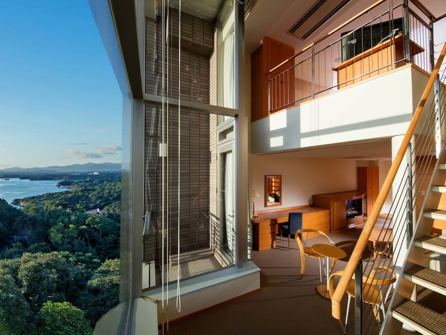 Un interior modern amb un balcó, una escala i vista a una zona boscosa.