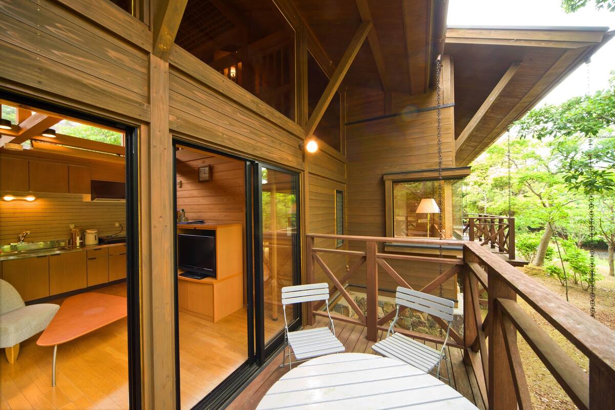 Une terrasse en bois avec une table et des chaises, un coin cuisine équipé d’un évier et de placards, et une vue sur la verdure environnante.