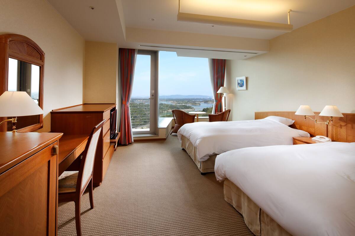 Une chambre d’hôtel avec deux lits, un bureau, une chaise, une petite table et une vue sur l’extérieur.