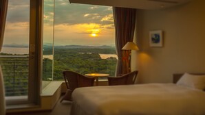 Een hotelkamer met een bed, een klein tafeltje en een stoel. Een groot raam biedt uitzicht op een landschap met bergen en een waterpartij.