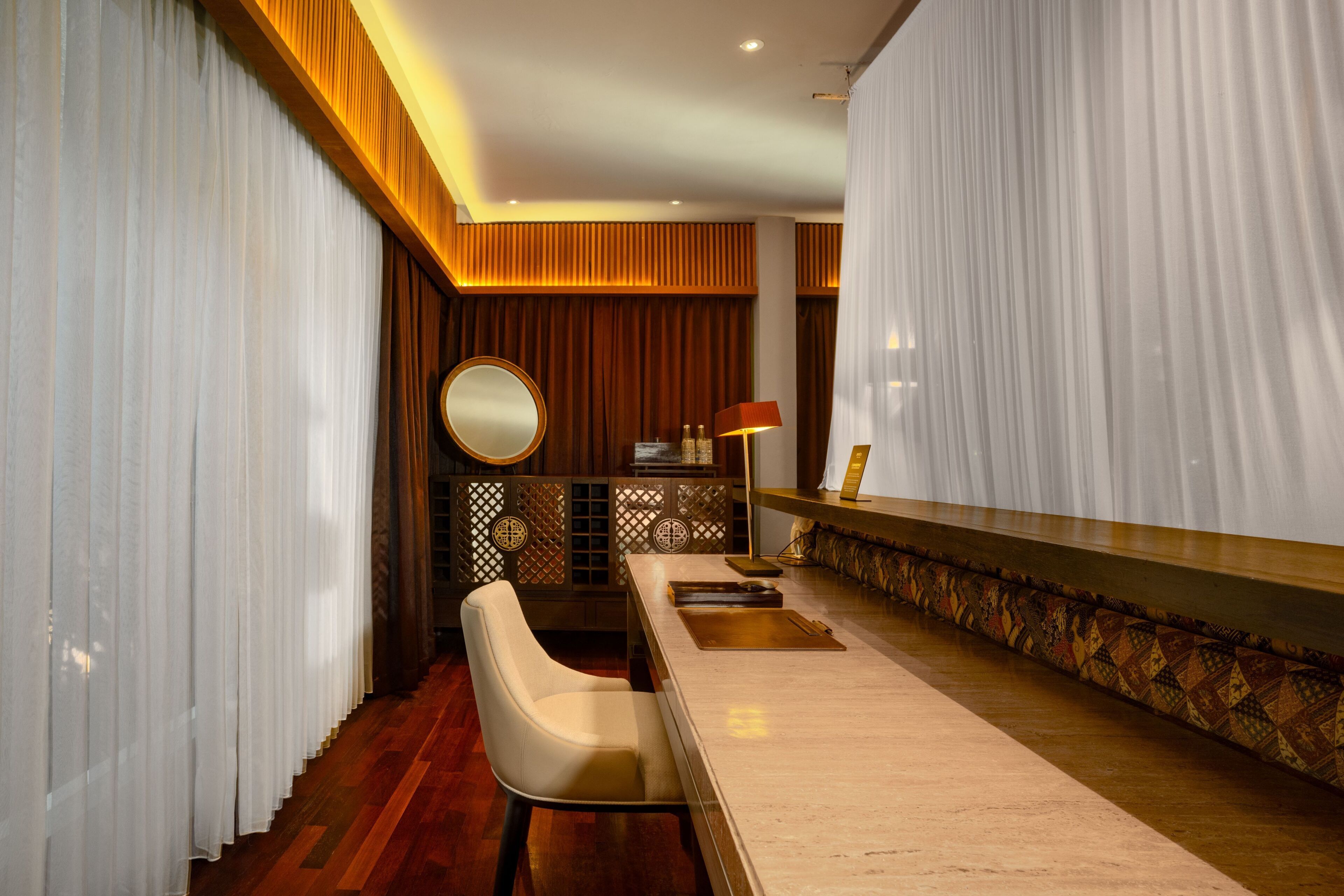 A hotel room with a wooden floor, a white chair, a round mirror, a bar area with a patterned rug, and a decorative wall panel.