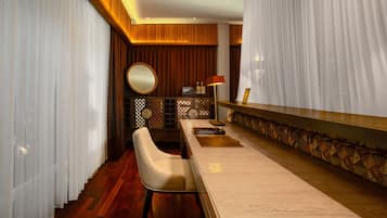 A hotel room with a wooden floor, a white chair, a round mirror, a bar area with a patterned rug, and a decorative wall panel.