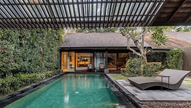 A swimming pool with a lounge area, a covered patio, and a house with a visible interior.