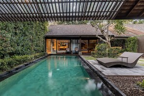 Ein Schwimmbecken mit Liegebereich, einer überdachten Terrasse und einem Haus, dessen Innenraum sichtbar ist.