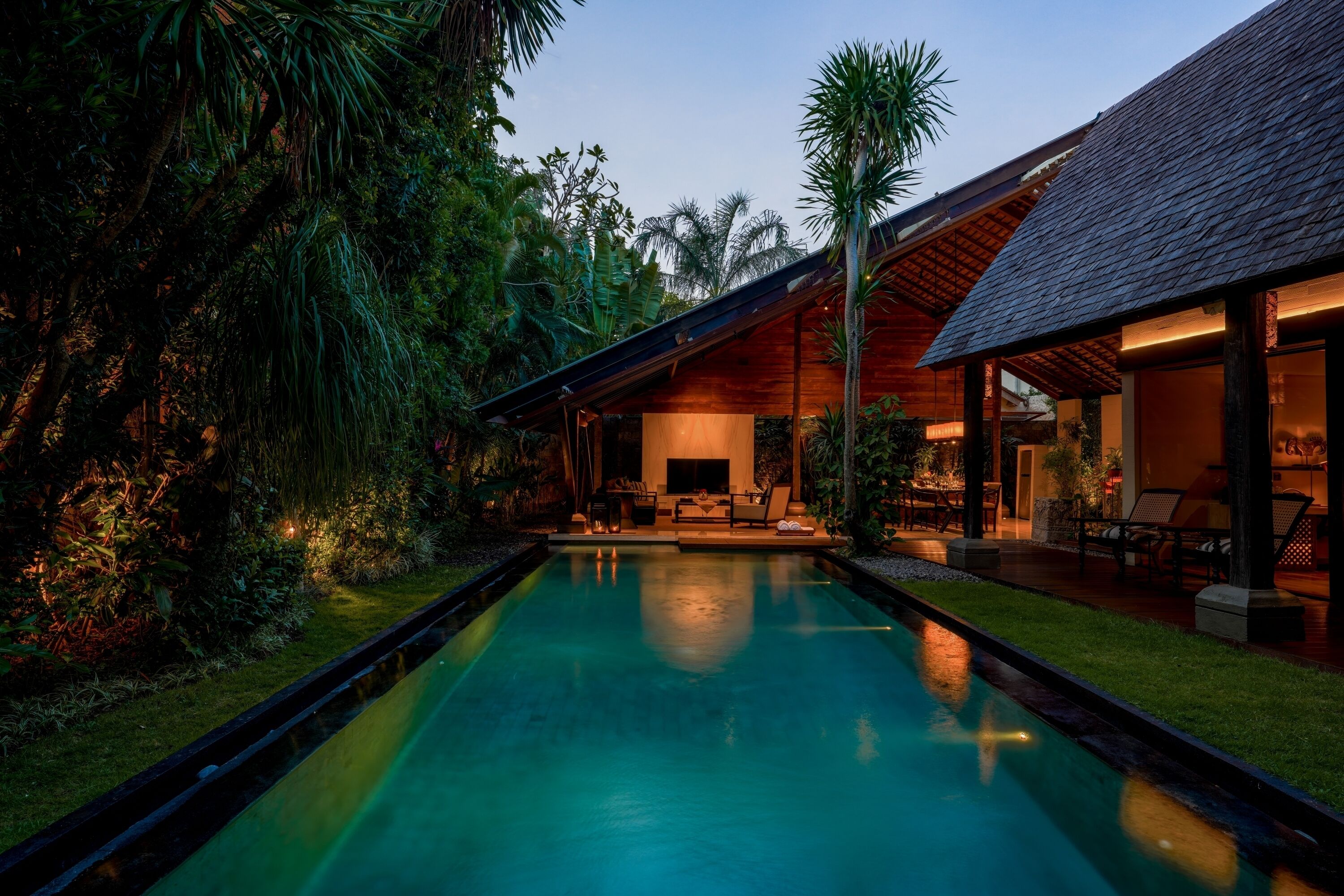 A pool with a wooden deck, surrounded by lush greenery and a modern house with large windows.