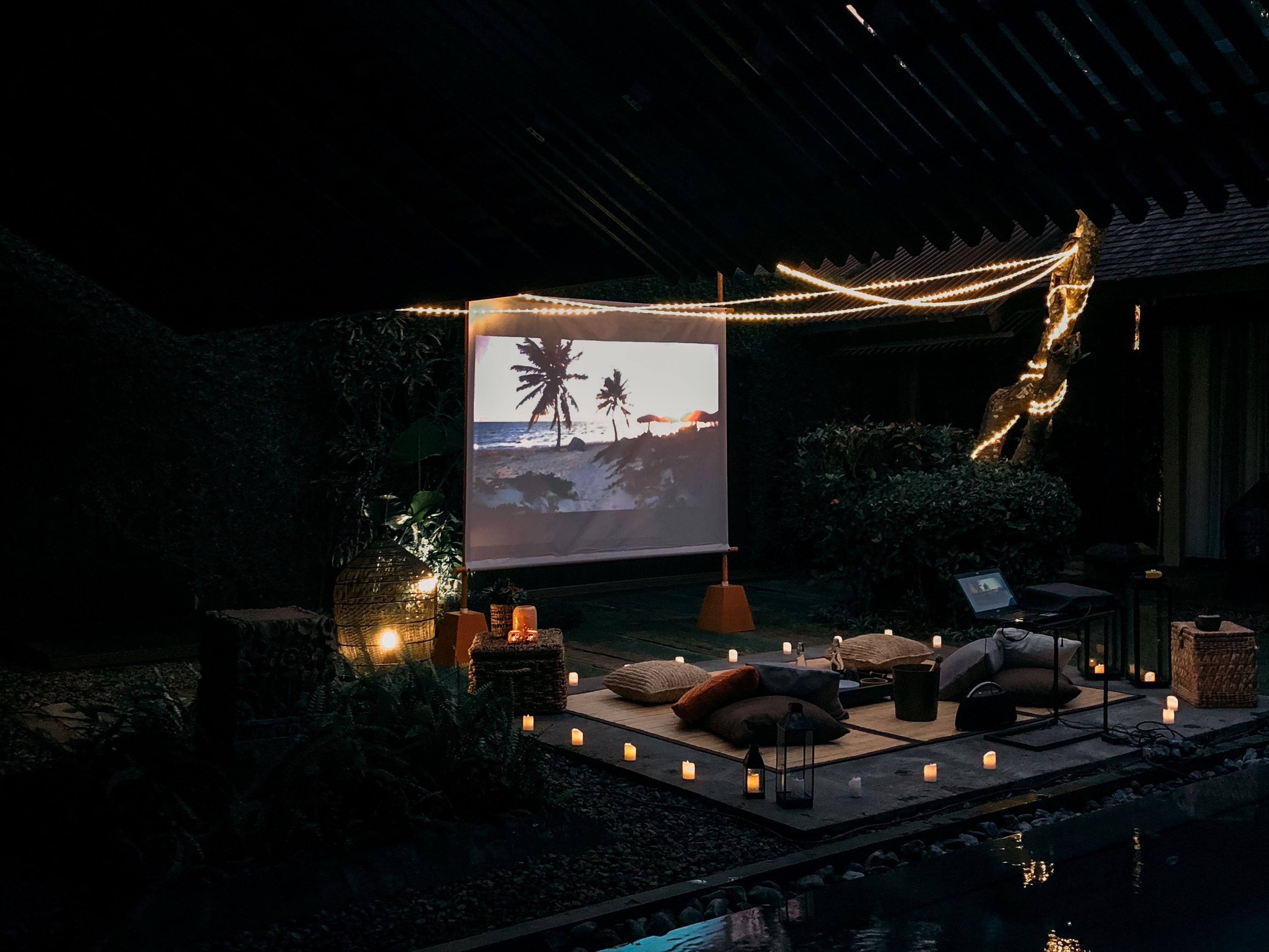 A serene outdoor seating area with a projector displaying a beach scene, surrounded by candles and ambient lighting.