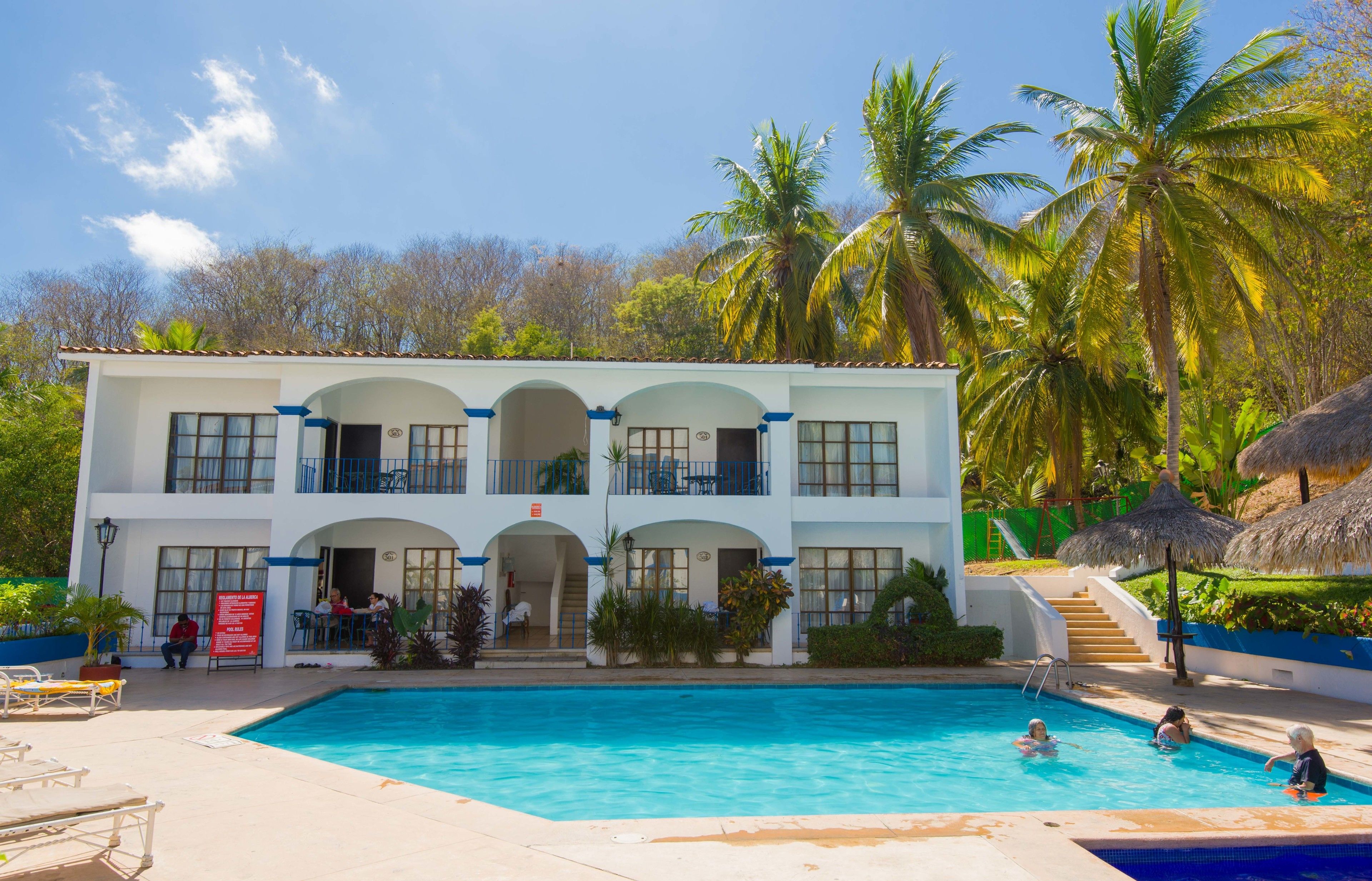 A two-story hotel with a swimming pool, outdoor seating, and palm trees.