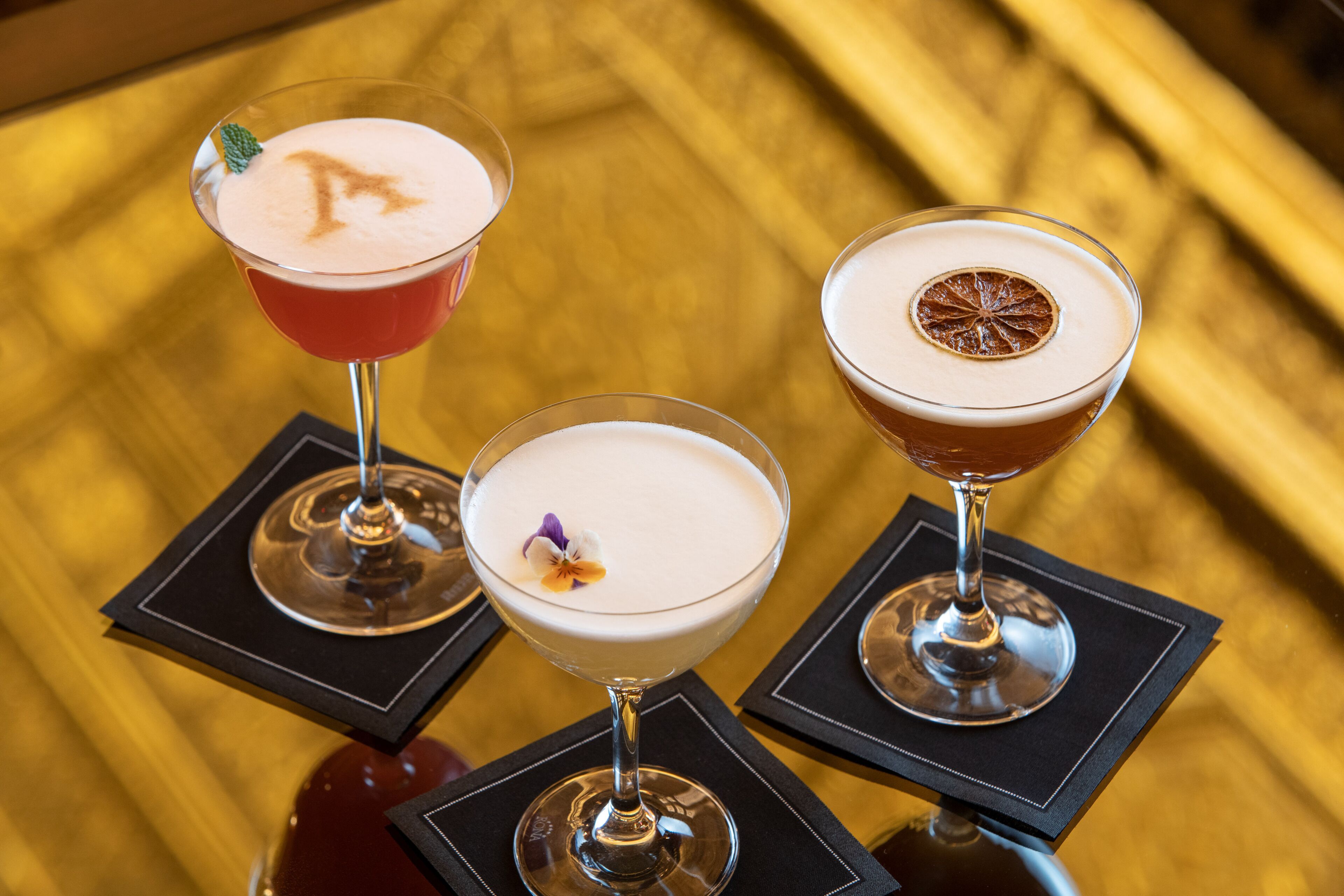 Tres cócteles sobre posavasos negros en una superficie reflectante.