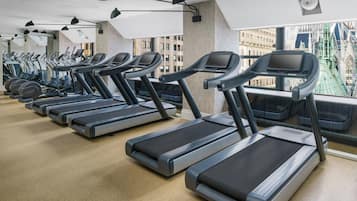 Un gimnasio con cintas de correr alineadas en filas, amplias ventanas con vistas a edificios y un techo alto con vigas a la vista.