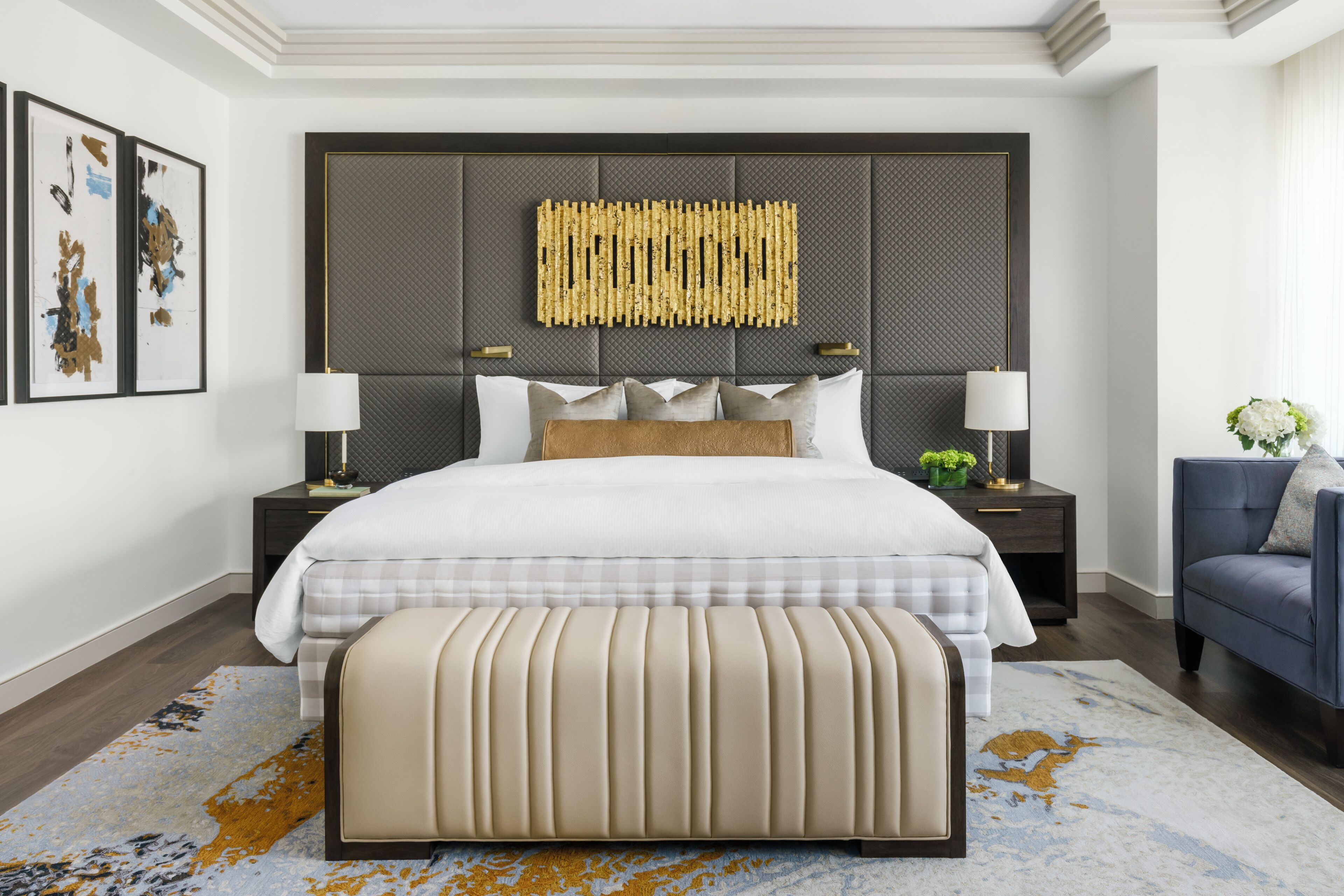 Una habitación de hotel moderna con una cama grande, un cabecero con panel texturizado, dos mesitas de noche con lámparas, un sillón azul y un elemento decorativo en la pared.