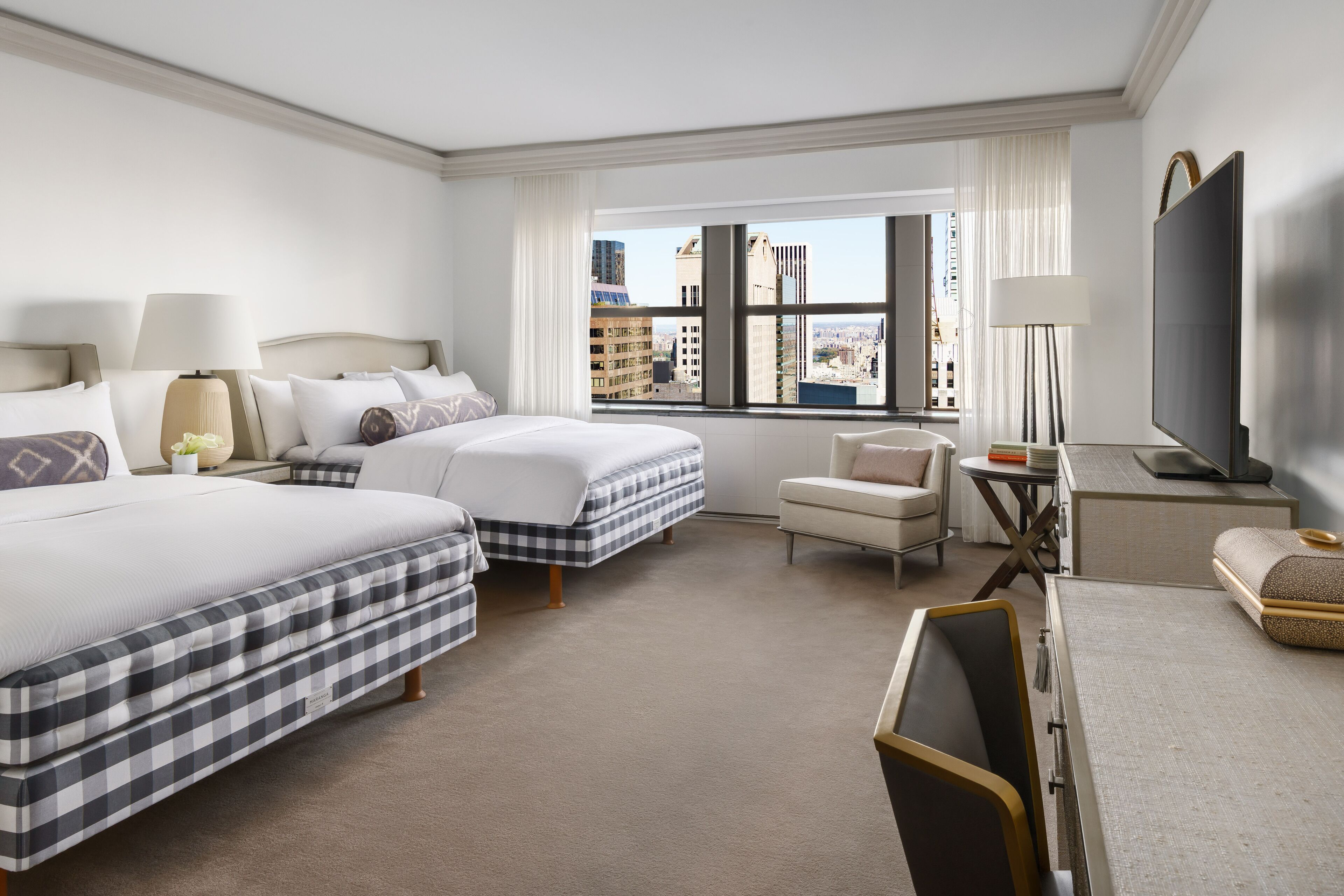A hotel room with two beds, a desk, a chair, a TV, and a large window with a city view.
