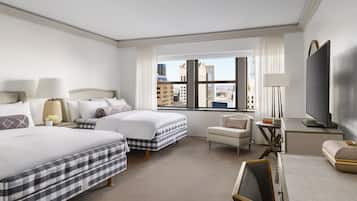 A hotel room with two beds, a desk, a chair, a TV, and a large window with a city view.