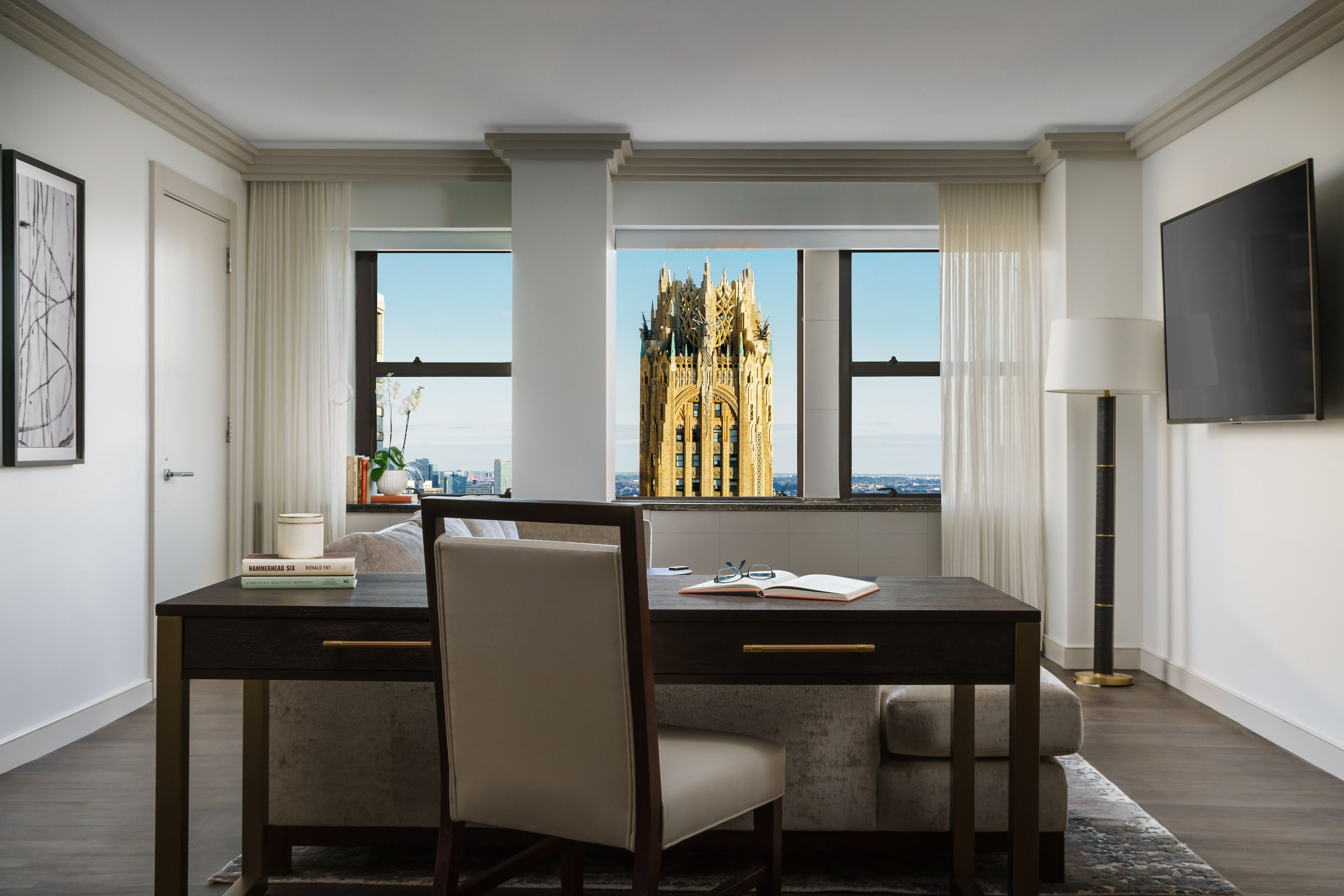 A modern hotel room with a large window offering a view of a distinctive building. There is a desk with a chair, a flat-screen TV, and a floor lamp.