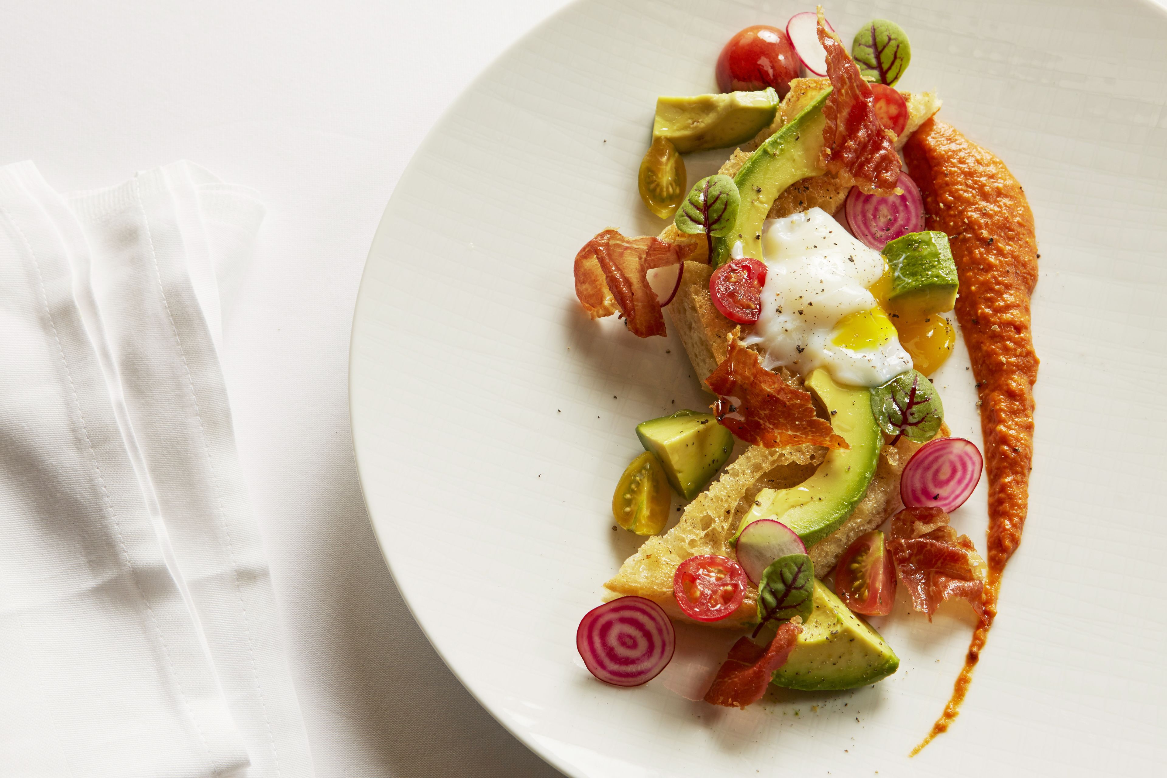 A white plate with a dish of avocado, tomatoes, radishes, and bacon.
