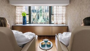 Una habitación con dos sillones reclinables orientados hacia una ventana con vistas a un edificio. Hay una mesita con dos tazas y un florero con flores.