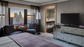 Habitación de hotel con una cama grande, dos sillones, un televisor, una pequeña mesa con un florero y vista a un paisaje urbano a través de la ventana.