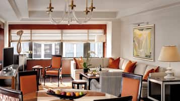 A modern hotel room with a dining area, a sofa set, a chandelier, and a large window with blinds.