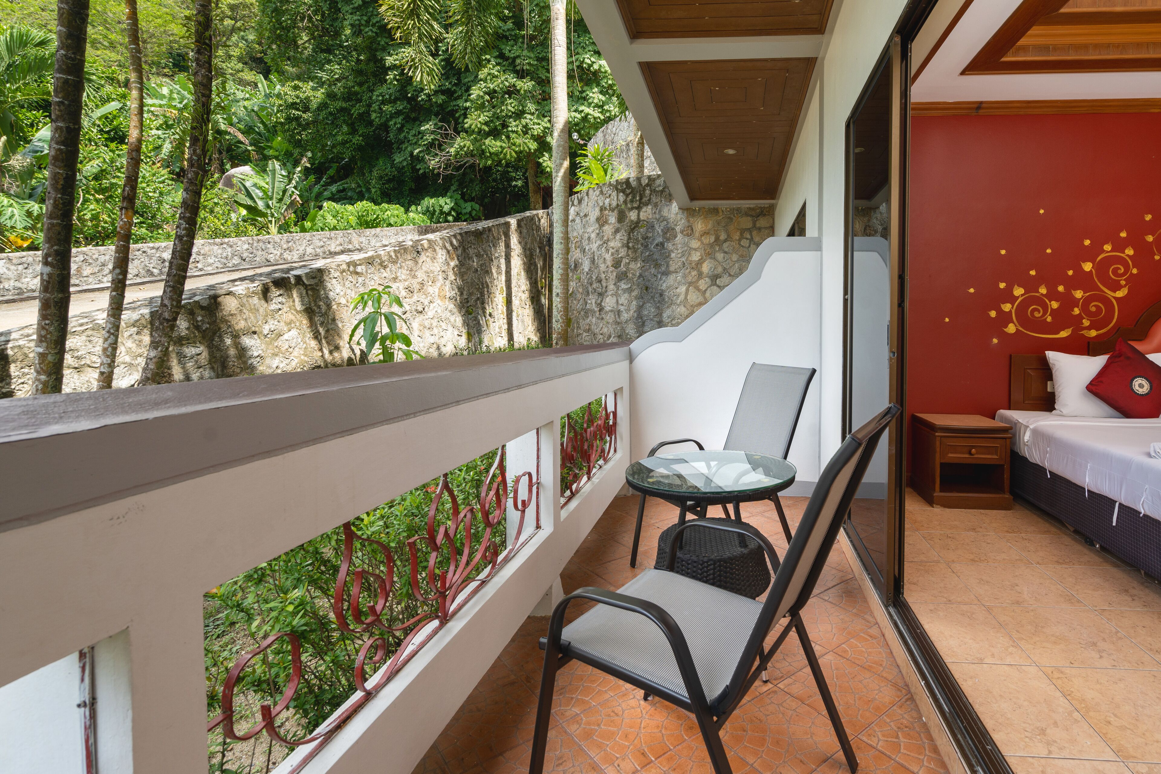 A balcony with a glass table and chairs, a bed with a red wall and decorative elements, and a view of greenery and a stone wall.